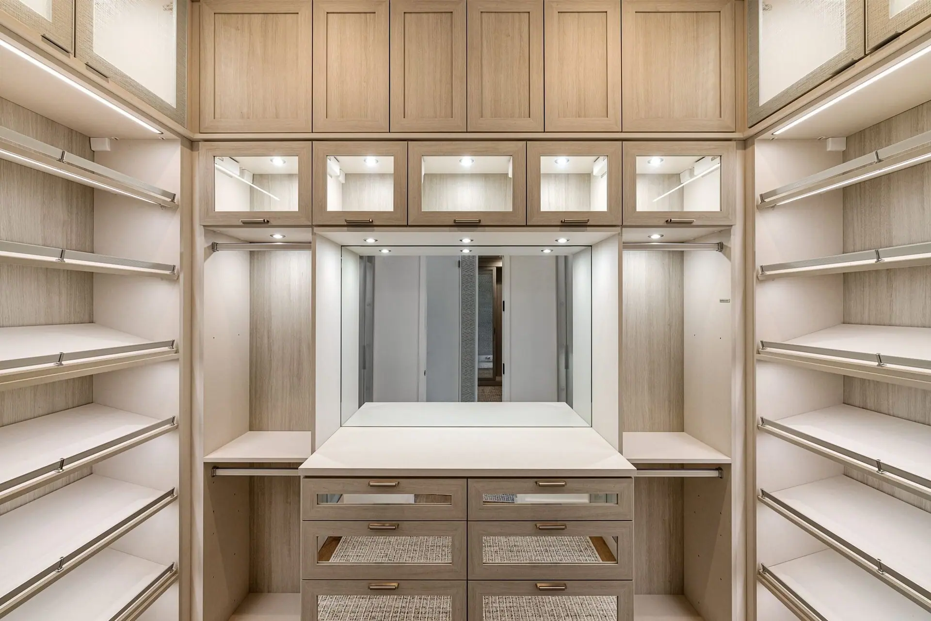 Pale oak walk-in wardrobe with lit shelves, glass-front cabinets and a central dresser with mirror